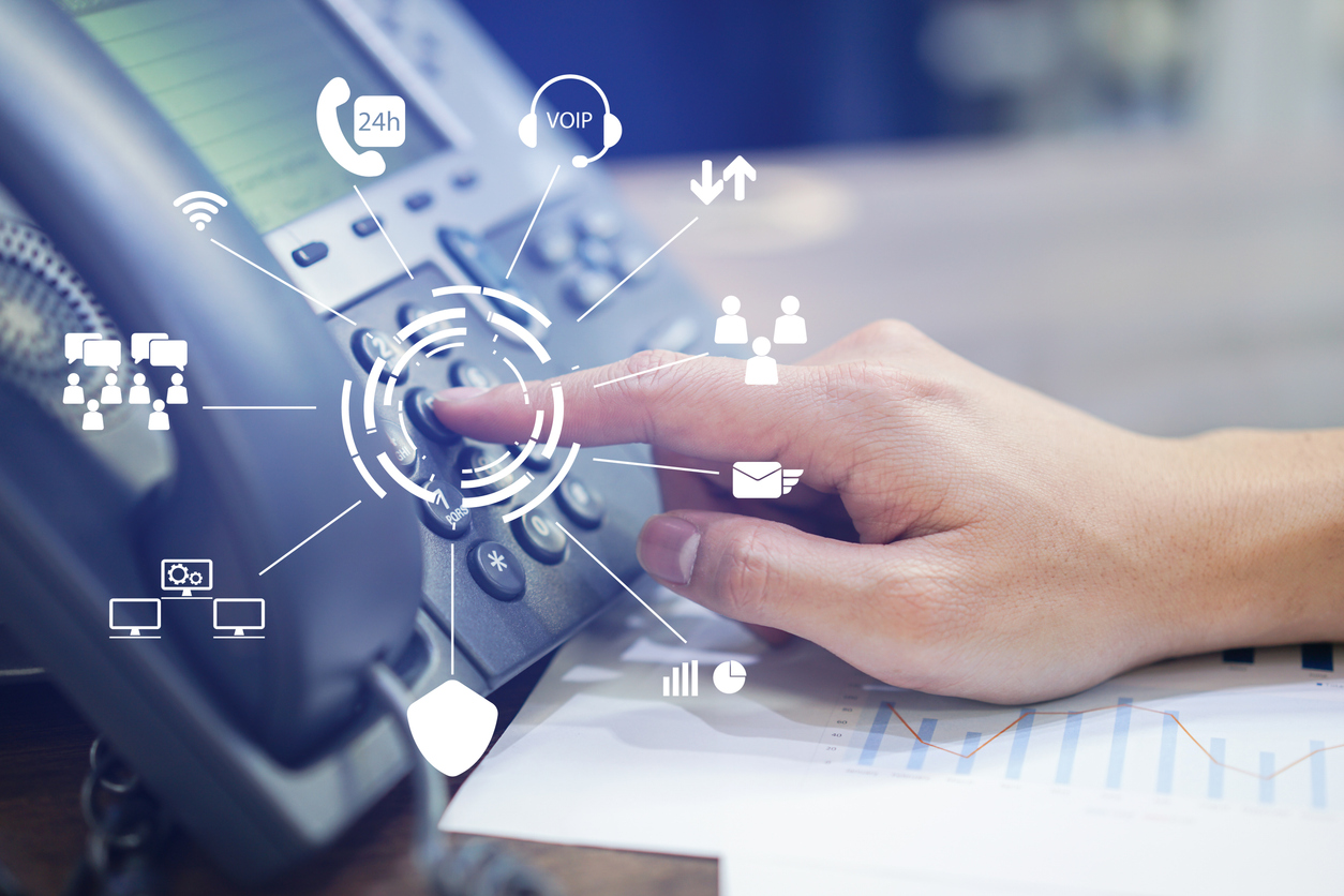 A hand pressing a button on a desk phone. Surrounding the button are various overlay icons representing communication concepts such as VOIP, email, 24-hour support, charts, security, and user connections. A graph is visible on a paper beside the phone.