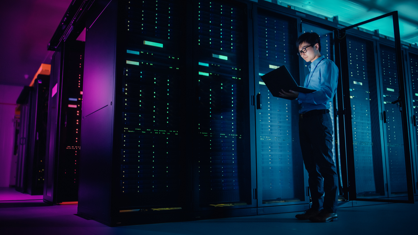 A person in a server room stands next to a row of illuminated servers, holding and examining a laptop. The dimly lit environment features colorful lighting, creating a technological and futuristic ambiance.