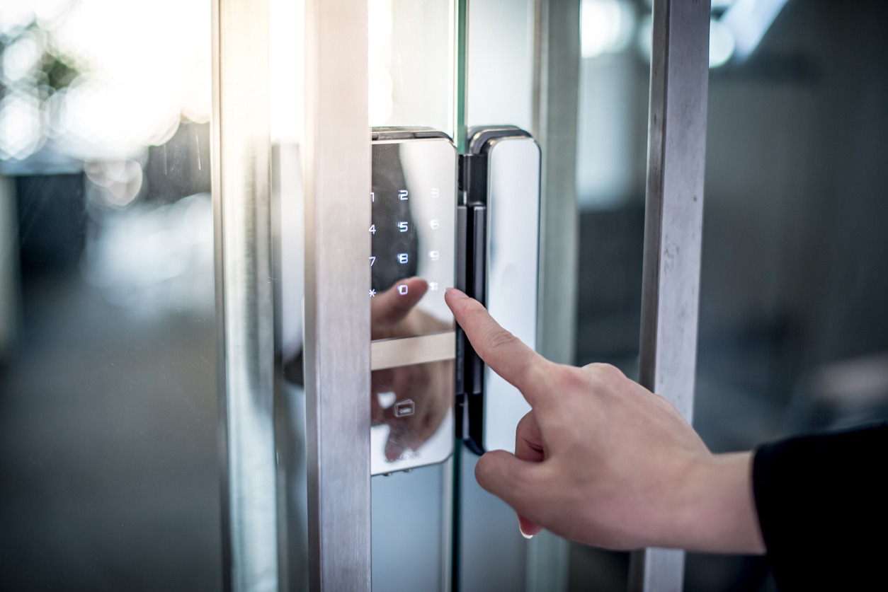 A close-up of a person's hand pressing a key on a digital keypad lock mounted on a glass door. The person's index finger is extended and touching one of the keys, suggesting they are entering a code to unlock the door.