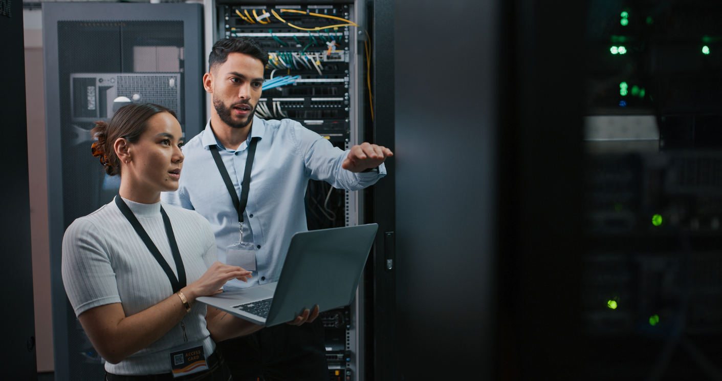 Two IT professionals work in a server room. The woman holds and types on a laptop while the man gestures towards a server rack. Both wear lanyards and ID badges. Various network cables and server equipment are visible in the background.