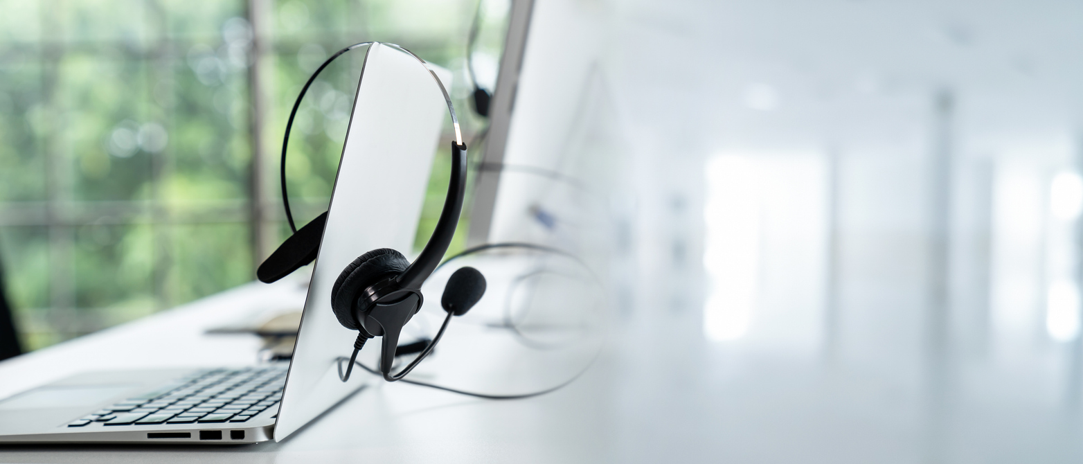 Close-up of a laptop with a headset resting on it, positioned on a white desk. The background is blurred, showcasing large windows with a view of green foliage, creating a calm and professional workspace atmosphere.