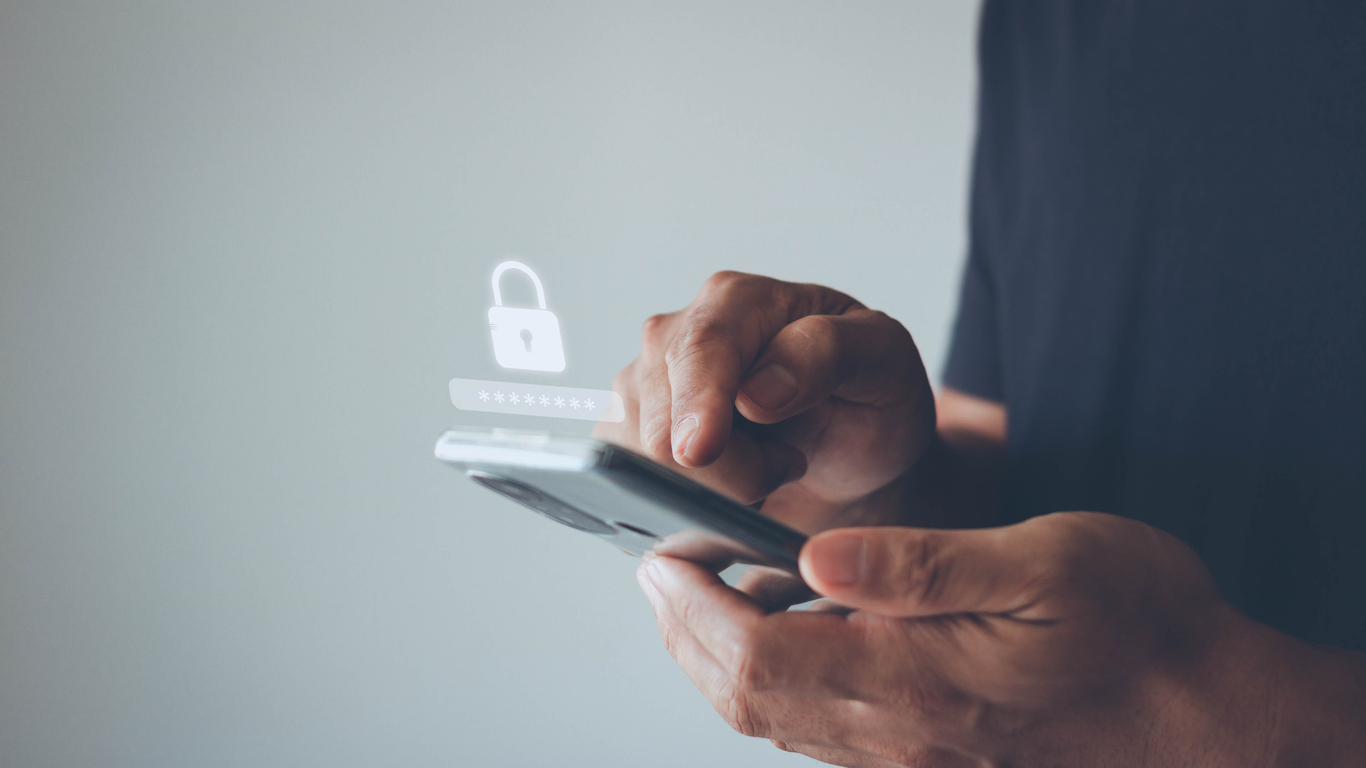 A person holding a smartphone with one hand and using the other hand to touch the screen. A digital image of a lock appears in mid-air above the phone, symbolizing security or password protection. The background is plain and blurred, focusing on the hands and phone.