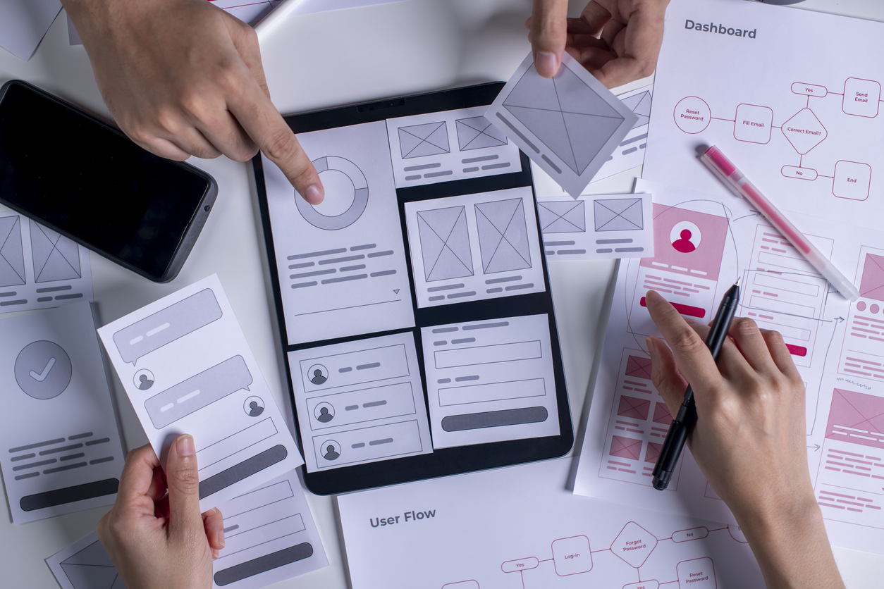 Hands of several people working collaboratively on UX/UI design layouts, reviewing wireframes and prototypes using printed papers, a tablet, and mobile devices. Various design elements and user flow diagrams are spread across the table.