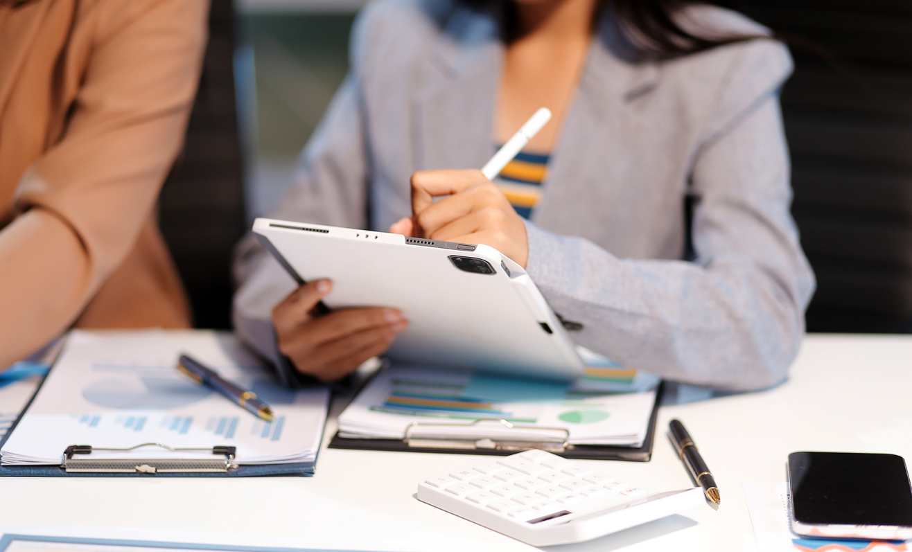 A person wearing a grey blazer uses a stylus on a tablet while sitting at a desk. The desk has a calculator, a clipboard with charts, and a pen. Another person in a brown jacket sits next to them.