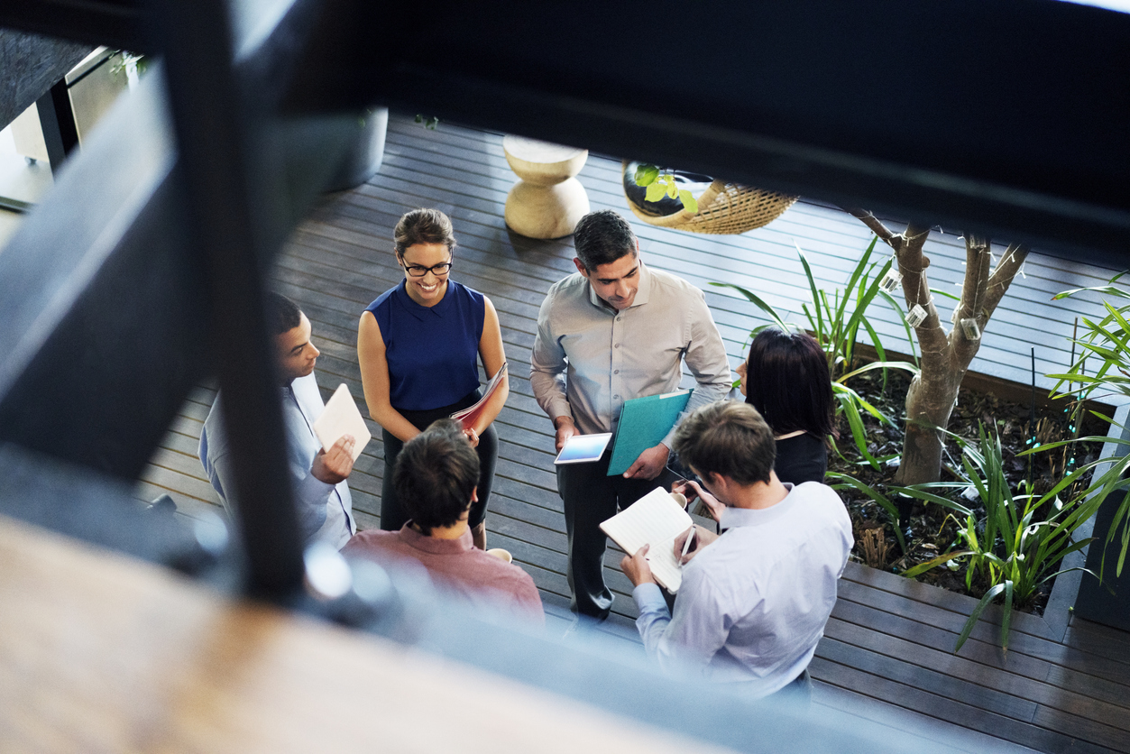 A group of six people, gathered in a casual business setting, are standing in a circle with notebooks and folders. They appear to be having a discussion. The surrounding area features wooden flooring, potted plants, and ample natural light.