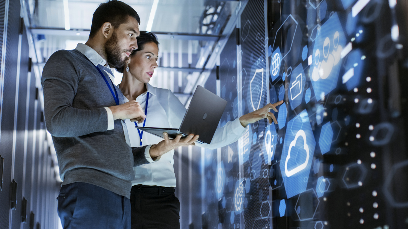 Two people stand in a futuristic server room with blue digital icons projected on a transparent screen. One is holding a laptop, while both point and discuss the data or interface on the screen. They appear engaged and focused on their work.