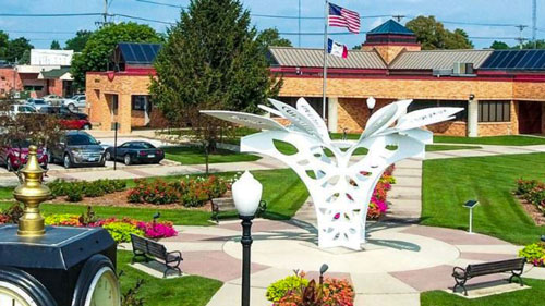 A modern white sculpture stands in a park surrounded by benches and greenery. An American and another flag are visible. A building with a red roof is in the background, and several cars are parked nearby.