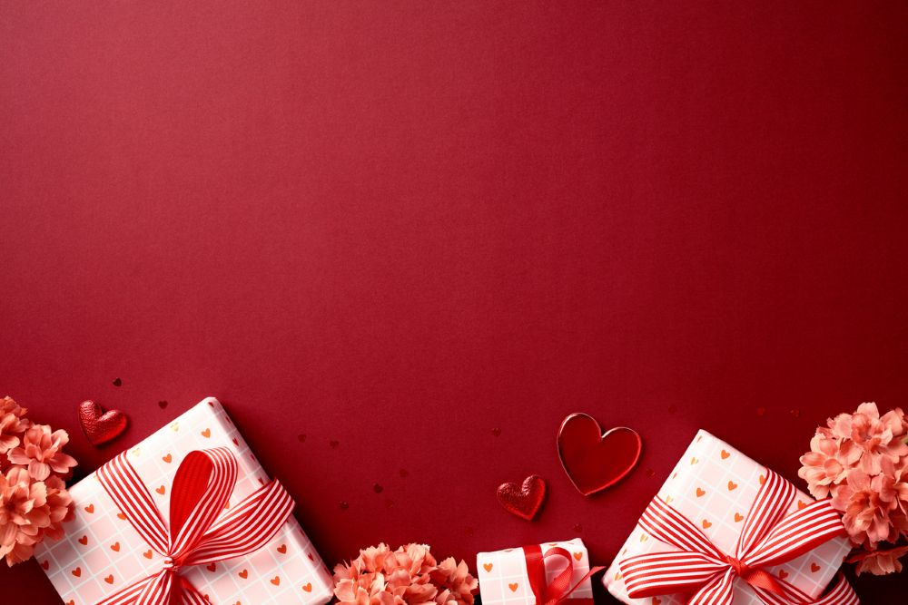 Red background with wrapped gifts and red ribbons in the bottom left corner. Pink flowers and heart decorations are scattered around. Ample space in the upper area.