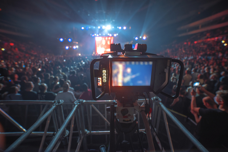 Close-up of a video camera recording a lively concert in a crowded arena. The stage is brightly lit in the distance, while the audience is mostly in shadow, showing a sea of people enjoying the performance.
