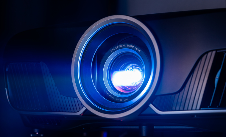 Close-up of a modern projector lens emitting a bright blue light. The surrounding casing is sleek and black with subtle ridges. The image captures the lens in a dark environment, emphasizing the vividness of the blue light.