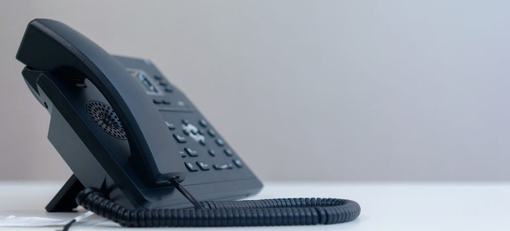 A black office desk phone with a corded handset sits on a white surface against a plain, light background—ideal for any phone promotion. The phone’s buttons and display screen are visible, and the coiled cord rests loosely in front.